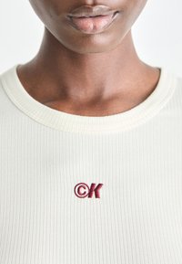 Ribbed light cream top featuring an embroidered dark red "CK" logo on the chest. Smooth texture with a round neckline.