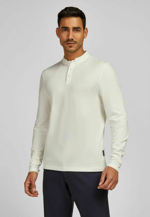 Langärmliges, cremefarbenes Henley-Shirt mit einem Drei-Knopf-Verschluss, weichem Material und dezentem LogoStick. Kombiniert mit dunklen Hosen.