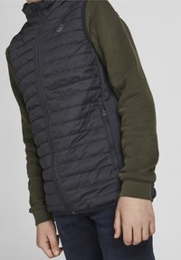 Boy wearing a dark quilted vest over an olive green long-sleeve shirt and dark pants, standing with hands relaxed by sides.