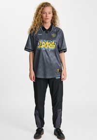 Szary, oversize'owy sweter z grafika "LOGOS" w żółtym i niebieskim, zestawiony z czarnymi, zwężającymi się spodniami z szarymi akcentami i ściągaczami na kostkach.