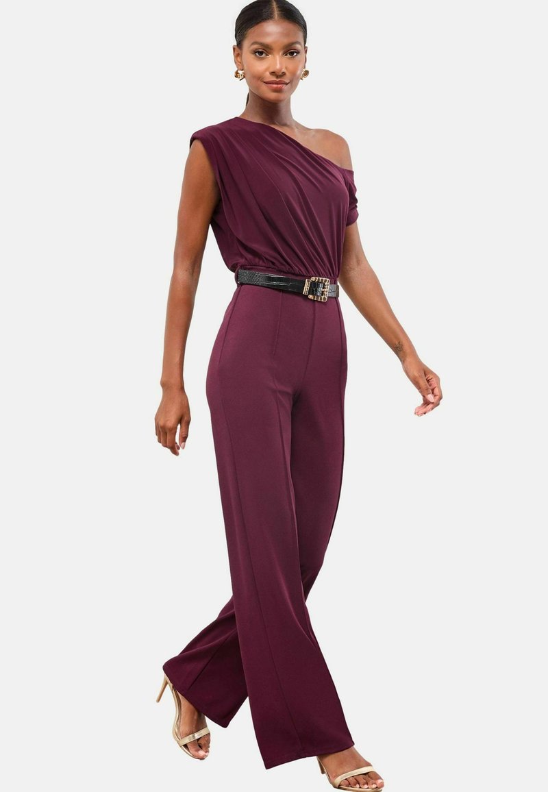 Eine Frau trägt einen ärmellosen lila Jumpsuit mit einem einseitigen Drapé, einem schwarzen Gürtel und beigen High Heels und geht selbstbewusst.
