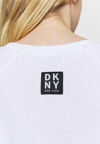 Bílá mikina s černým čtvercovým logem, na kterém je bílý text "DKNY NEW YORK", umístěným na zadním výstřihu.