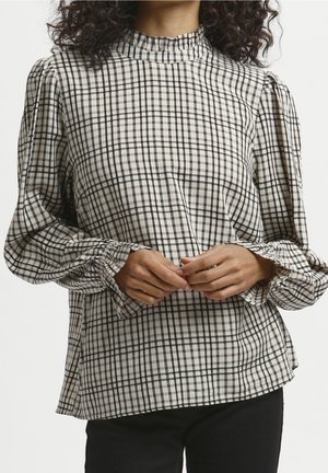 Donna che indossa una camicetta a maniche lunghe a scacchi bianchi e neri con polsini arricciati e collo alto, con le mani giunte davanti.