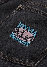 Ύφασμα μαύρου τζιν με κεντημένο λογότυπο σε ανοιχτό μπλε και ροζ, που απεικονίζει μια υδρόγειο σφαίρα και το κείμενο "RIVIA PROJECTS." Ορατές ραφές λεπτομερειών.