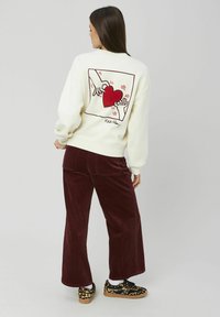 Sweat-shirt crème avec un graphique de cœur rouge et un design de mains au dos. Associé à un pantalon en velours côtelé bourgogne et des chaussures à motifs.