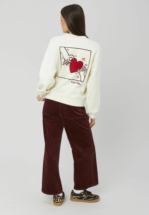 Sweat-shirt crème avec un graphique de cœur rouge et un design de mains au dos. Associé à un pantalon en velours côtelé bourgogne et des chaussures à motifs.