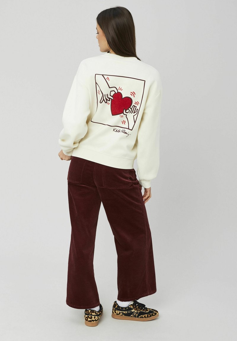 Sweat-shirt crème avec un graphique de cœur rouge et un design de mains au dos. Associé à un pantalon en velours côtelé bourgogne et des chaussures à motifs.