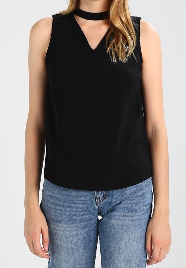 Femme portant un haut noir sans manches à col en V et col choker, associé à un jean bleu, debout devant un fond uni.