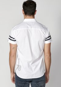 Camisa blanca de manga corta con rayas negras en las mangas, un dobladillo curvado y un estampado gráfico con el número "78" en la parte posterior, cerca de la costura lateral.