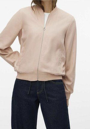 Woman wearing light pink zip-up bomber jacket and dark blue jeans with hands in pockets against white background.
