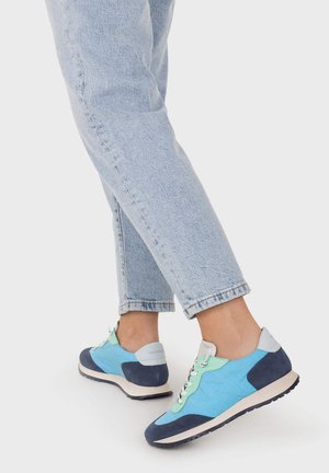 Personne portant un jean bleu clair et des baskets bleu-vert avec des lacets blancs, debout sur un fond blanc.