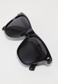 Glossy black square-frame sunglasses with dark tinted lenses placed on a white surface, folded with visible temples and a small logo detail.