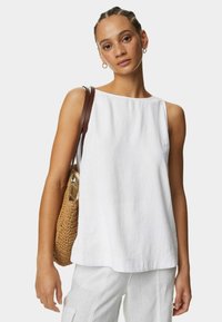 Haut sans manches blanc en lin avec un col rond. La modèle porte un sac fourre-tout tressé marron. Coupe simple et décontractée sans motifs.