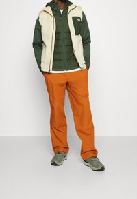 Uomo con le mani in tasca, che indossa una felpa verde con zip, un gilet senza maniche color crema, pantaloni arancioni e sneakers verdi, in piedi su un pavimento grigio.