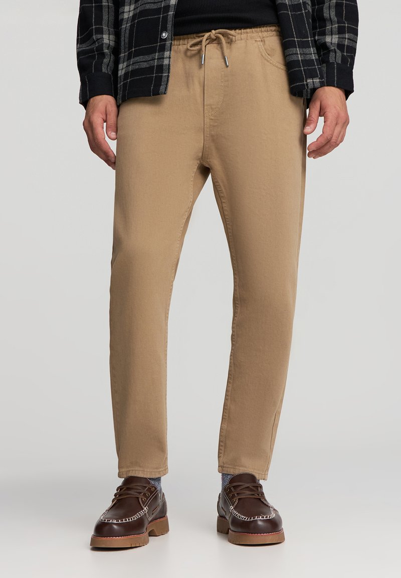 Jasnobrązowe spodnie o zwężanym kroju, z elastycznym paskiem i sznurkiem do ściągania. Połączone z ciemniebrązowymi butami i koszulą w kratę.