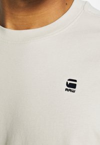 Světlobéžové bavlněné tričko s kulatým výstřihem. Na horní levé straně se nachází malé černé logo "RAW". Hladká textura, jednoduchý design.