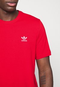 Červené bavlněné tričko s kulatým výstřihem a krátkými rukávy; má malé bílé logo Adidas na levé části hrudníku.