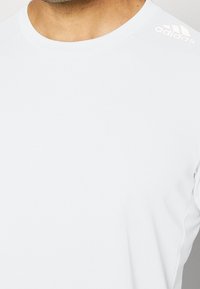 Camiseta deportiva de manga corta blanca con cuello redondo. Presenta una tela texturizada y un sutil logo de "adidas" en el hombro izquierdo.