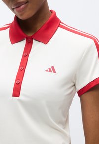 Polo blanc avec col et accents de manches rouges, doté d'une patte de boutonnage à trois boutons et d'un petit logo rouge sur le côté gauche.