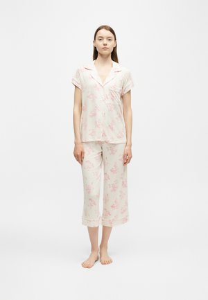 Femme debout pieds nus portant un haut blanc à manches courtes et un pantalon court à imprimé floral rose clair sur un fond uni.