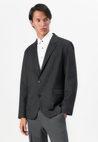 Junger Mann in einem dunklen Hahnentrittmuster-Blazer, weißem Hemd und grauen Hosen, der nach links schaut, vor einem schlichten weißen Hintergrund.
