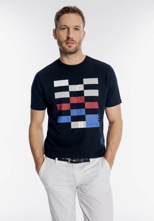 Mężczyzna o krótkich włosach i brodzie, ubrany w czarną koszulkę z czerwono-biało-niebieskim wzorem w kształcie bloków oraz białe spodnie, z rękami w kieszeniach.