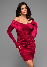 Abito rosso in velluto off-shoulder con design arricciato, maniche lunghe e silhouette aderente. Texture liscia e un leggero luccichio in tutto l'abito.