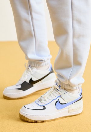 Białe i czarne sneakersy Nike Air Force 1 z niebieskimi akcentami noszone z jasnoszarymi spodenkami typu jogger na żółtej podłodze.