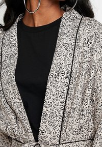Chaqueta ligera estampada en beige con diseño de hojas negras; presenta un ribete en contraste y un ajuste relajado. Llevada sobre una camiseta negra.
