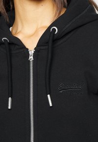 Lähikuva henkilöstä, jolla on päällä musta vetoketjullinen huppari, jossa on nimikirjaimet "SuperDry" brodeerattuna rinnassa ja metallipäiset narut.