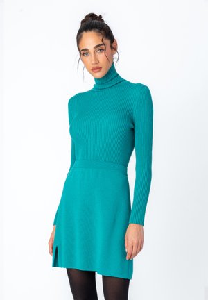 Ribbed teal turtleneck dress with long sleeves, fitted bodice, and flared skirt featuring a side slit. Smooth texture, slim silhouette.