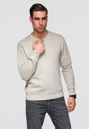 Jeune homme portant une chemise beige à manches longues et un jean foncé, touchant son col avec une expression neutre devant un fond uni.
