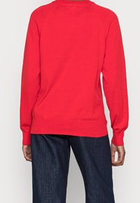 Czerwony sweter dzianinowy z długimi rękawami raglanowymi, okrągłym dekoltem oraz ściągaczami przy rękawach i na dole. Połączony z ciemnymi dżinsami.