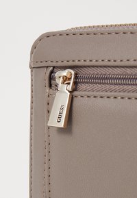 Billetera de piel sintética gris con cremallera y herrajes en tono dorado, que presenta una fina costura y una superficie texturizada. Logo de la marca visible en el tirador de la cremallera.