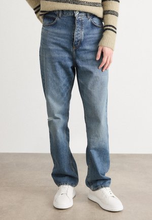 Blue denim jeans with a loose fit, featuring a front rip, standard pockets, and a button closure. Paired with white sneakers.
