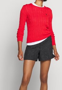 Röd kabelstickad tröja lager-på-lager över en vit T-shirt, kombinerad med svart- och vitrandiga shorts. Tröjan har ribbade muddar och fåll.