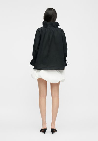 Γυναίκα που φοράει μαύρο τοπ με μακριά μανίκια, βολάν στο γιακά και τα μανίκια, κοντή λευκή φούστα με φουσκάλες και μαύρα mule με τακούνι, από πίσω.