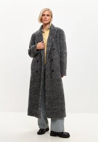 Dlouhý šedý kabát s texturou vyrobený z chlupatého materiálu, dvojitý knoflíkový zapínání, klopový límec a černé knoflíky. Stylizován přes neformální oblečení.