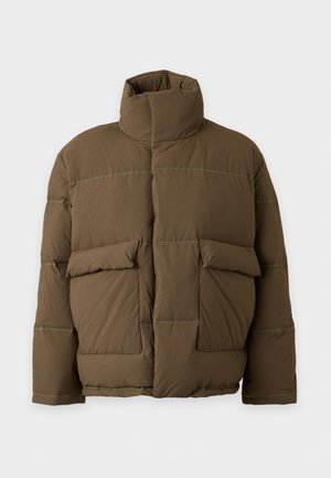 Un homme porte une veste matelassée marron, un polo rayé, un pantalon crème, des baskets marron et une casquette noire sur un fond uni.
