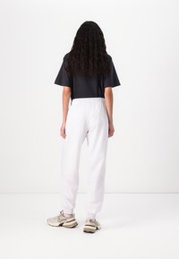 Μαύρο μπλουζάκι με κοντά μανίκια, λευκή φόρμα με ελαστική μέση και μπεζ αθλητικά παπούτσια. Τα σγουρά μαλλιά πέφτουν στην πλάτη, με ουδέτερο φόντο.