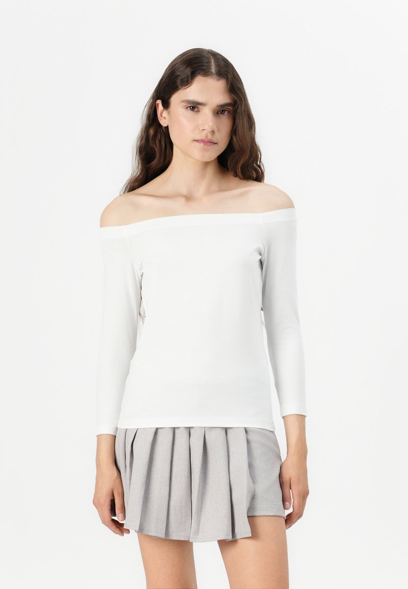 Top bianco off-shoulder con maniche lunghe, realizzato in un tessuto morbido. Caratterizzato da una trama liscia e una silhouette aderente. Abbinato a una gonna grigia plissettata.