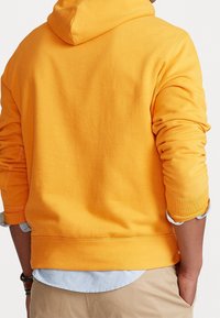 Gul bomullshuvjacka med långa ärmar och snodd i huvan. Har ribbade mudd och nederkant, med en lager av en ljusblå skjorta under.