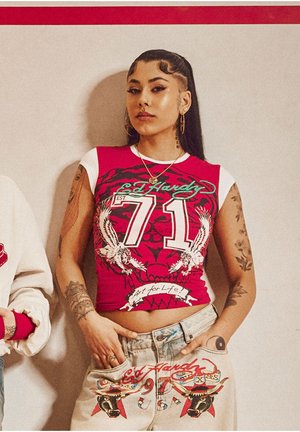 Young woman with long dark hair and tattoos wears a red and white graphic crop top and light denim pants with bold Ed Hardy designs.
