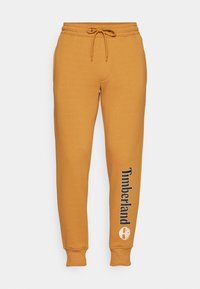 Pantalones de chándal color mostaza con cinturilla elástica y cordón ajustable, que presentan el texto y el logotipo "Timberland" en la pierna izquierda.