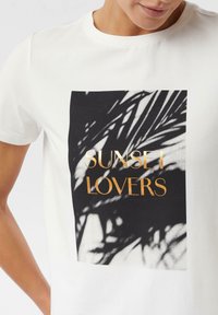 Biała bawełniana koszulka z czarno-białym nadrukiem palmowych liści oraz napisem "SUNSET LOVERS" w złotych literach.