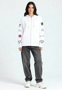 Felpa con cappuccio bianca con cerniera e varie grafiche in nero e a colori sulle maniche, abbinata a jeans grigi a taglio largo e sneaker bianche.