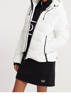 Personne portant une doudoune blanche avec des détails de fermeture éclair noirs et une robe noire avec un petit patch logo sur l'ourlet.