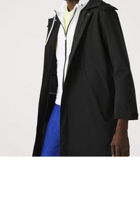 Černý voděodolný kabát se strukturovaným designem, s kapucí, dlouhými rukávy a bočními kapsami. Nosený přes bílou bundu na zip a žlutou košili.