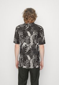 Lyhythihainen musta paita, jossa on valkoinen trooppisen lehden kuvio, kaareva helma ja rento istuvuus. Takakuva näyttää kauluksen ja tekstuurillisen kankaan.