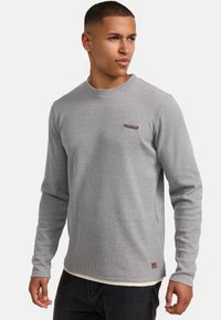 Grijze geribde langermige shirt met een ronde hals, gestructureerde stof en een klein bruin merklabel op de borst.